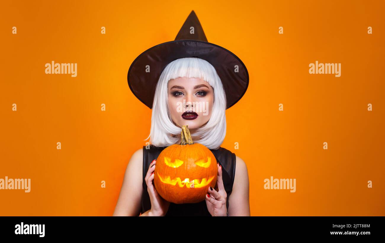 Beautiful sexy woman in witch hat and costume holding carved pumpkin on orange background. Stock Photo