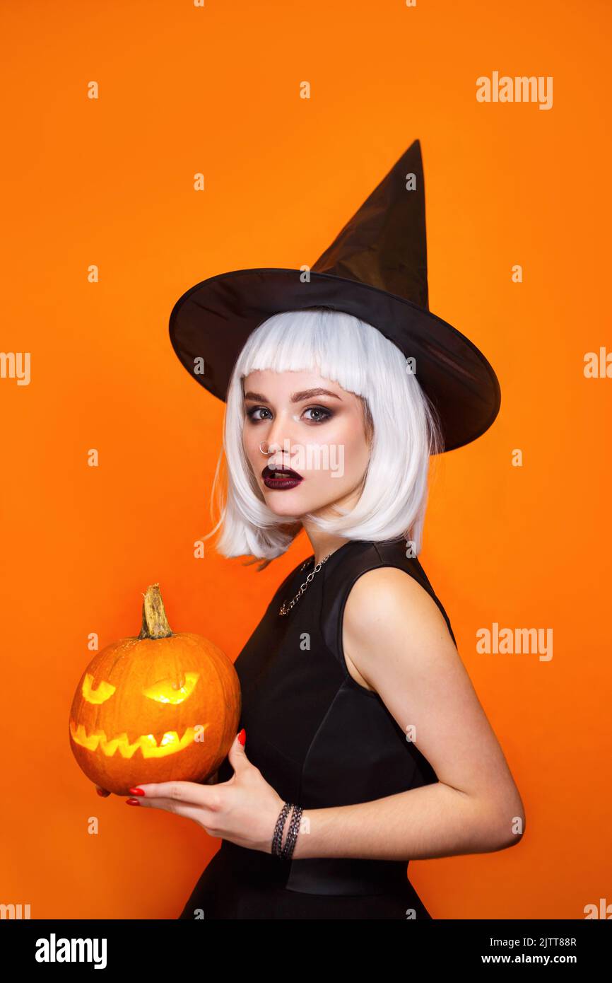 Beautiful sexy woman in witch hat and costume holding carved pumpkin on orange background. Stock Photo