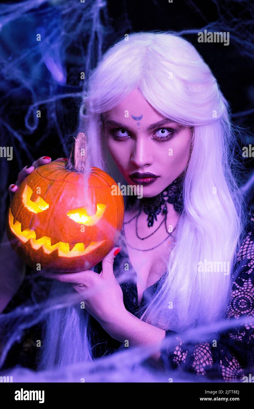 Blond woman witch with long white hair holding pumpkin. White lenses in the eyes. Halloween party. Stock Photo