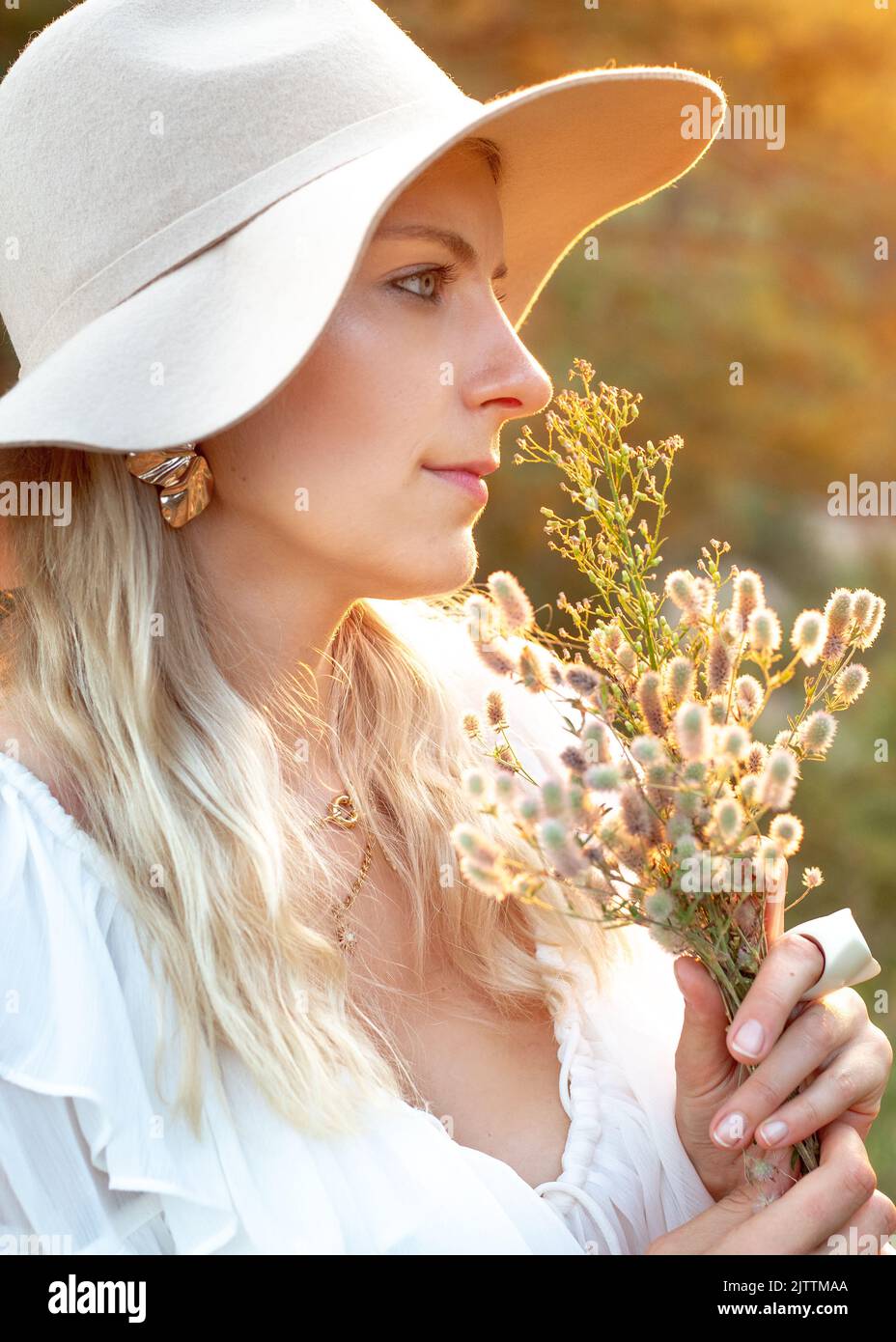Side view of young adorable woman wearing white dress, beige floppy hat, holding, sniffing at bouquet of wildflowers. Stock Photo