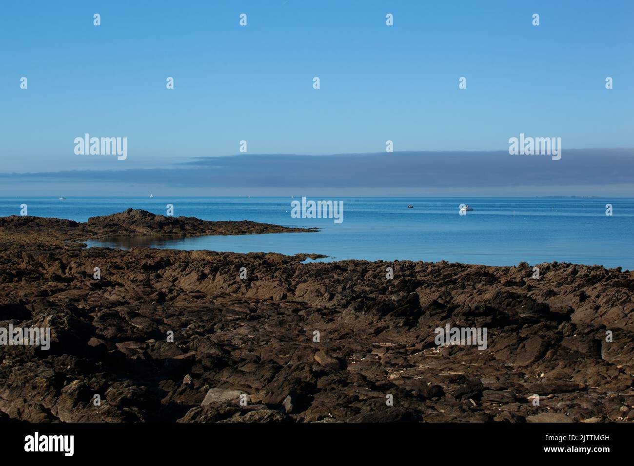 The carved rocks plunge into the blue sea. Offshore, boats sail on the calm water. High clouds block the horizon. Stock Photo