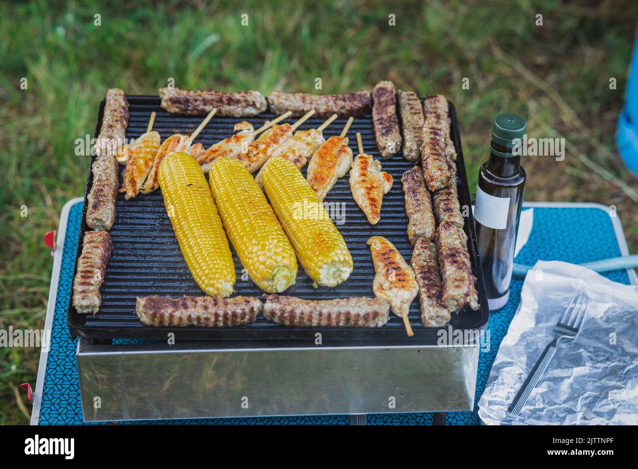 Delicious assortiment of meat and corn on a small gas cooker at a picnic outdoors. Tasty food being prepared on a table top grill. Stock Photo