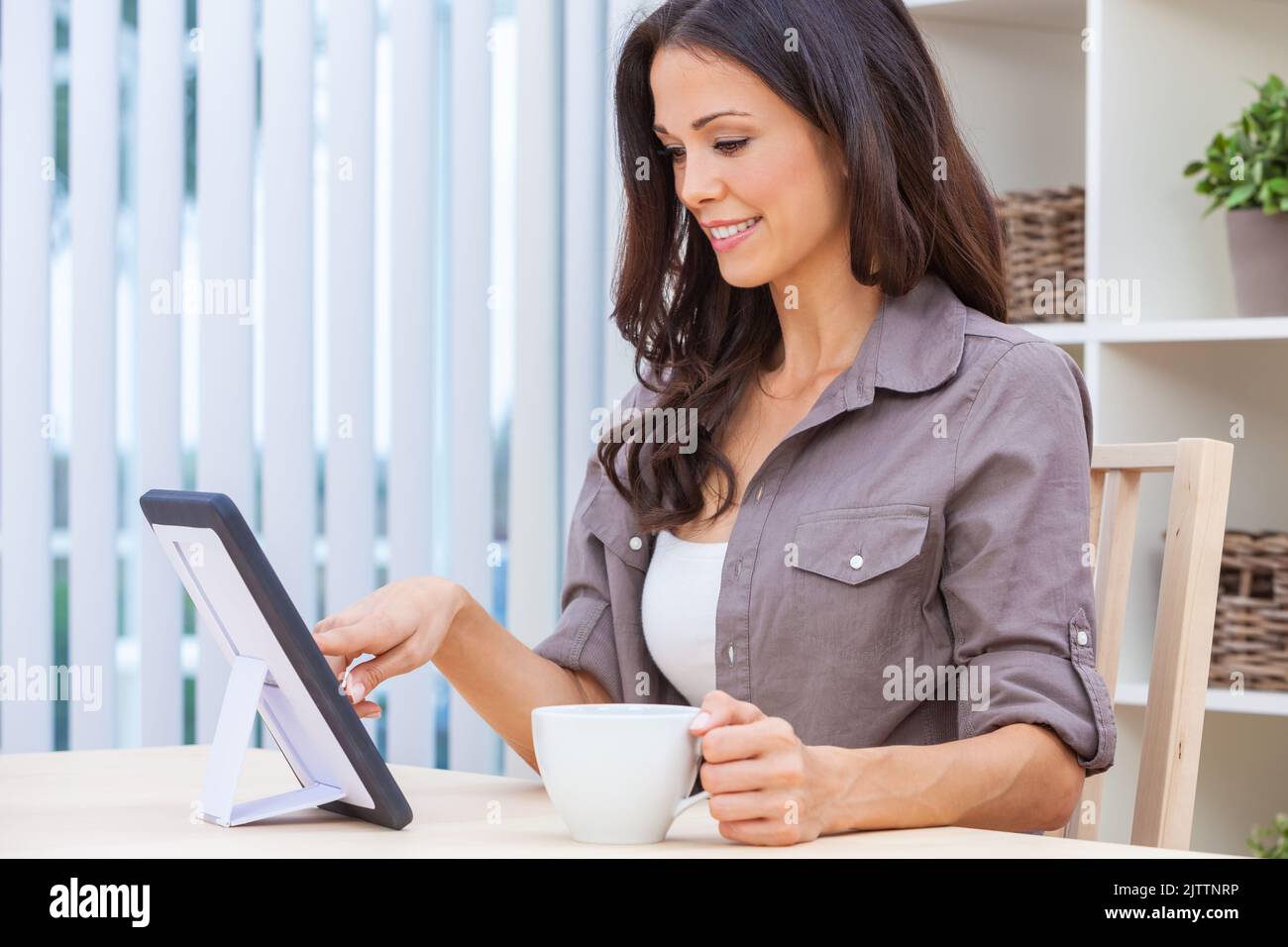 Beautiful girl young woman female at home using her tablet computer for social media, work or on line shopping smiling drinking a cup of tea or coffee Stock Photo
