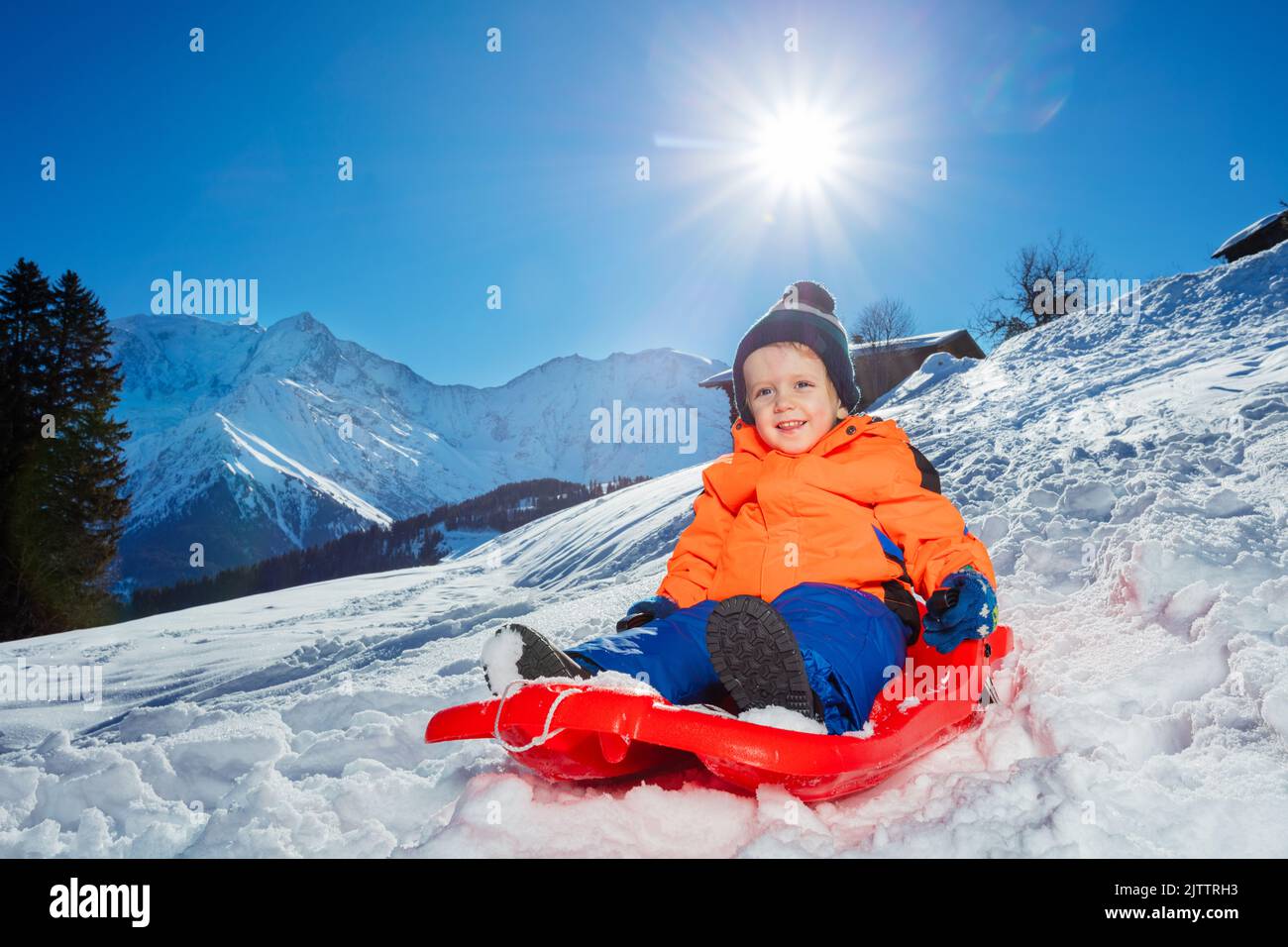 Cute little boy go downhill in the red sledge at mountains Stock Photo