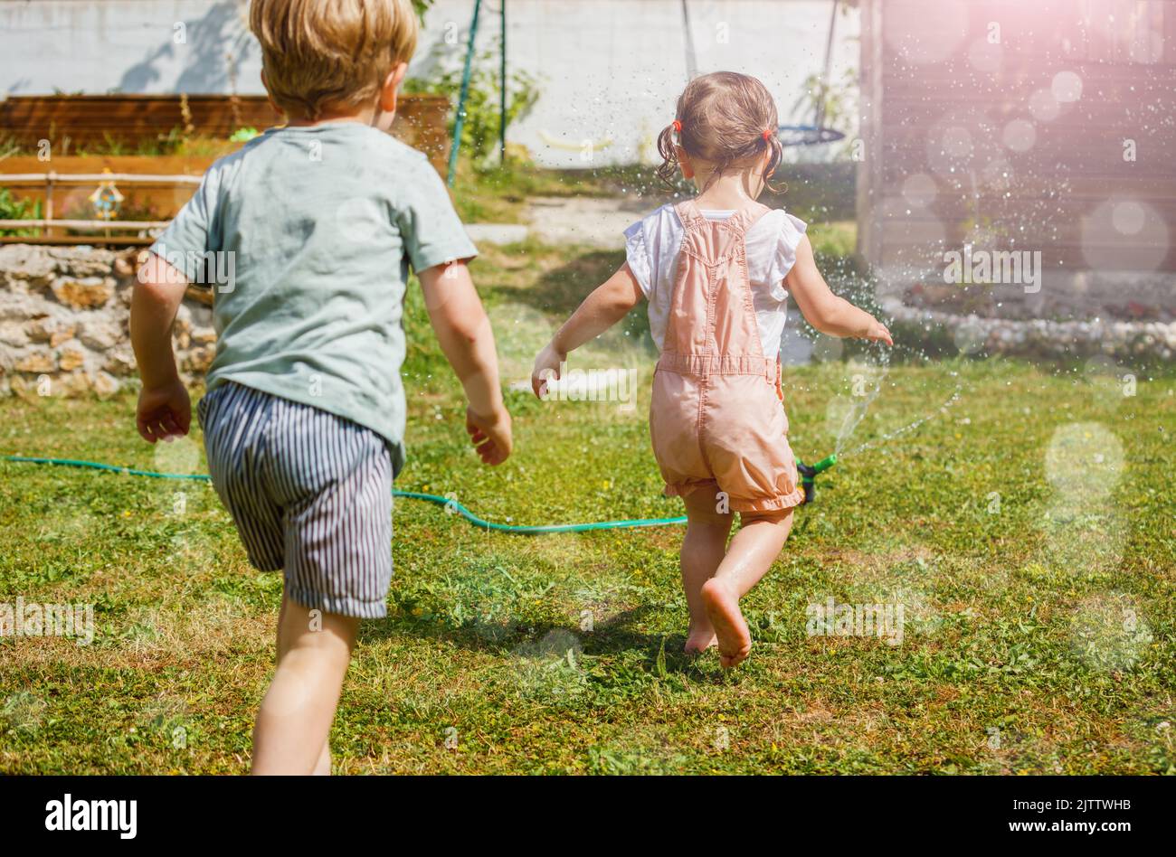Two happy little kids boy, girl run around water sprinkler Stock Photo