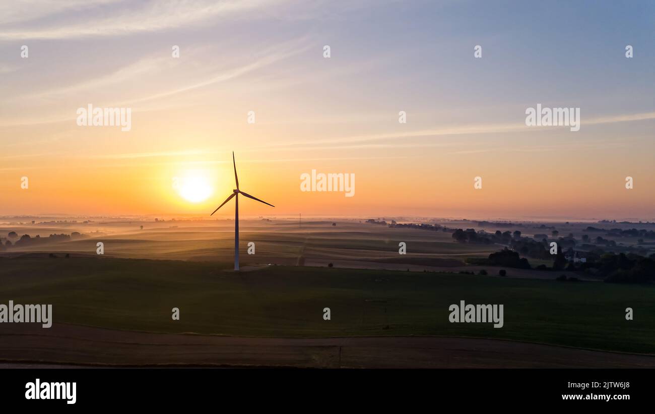 Bright orange rising sun over farmfields with silhouette of wind turbine and trees. Colorful sky. Environmentally friendly energy. Horizontal shot. High quality photo Stock Photo