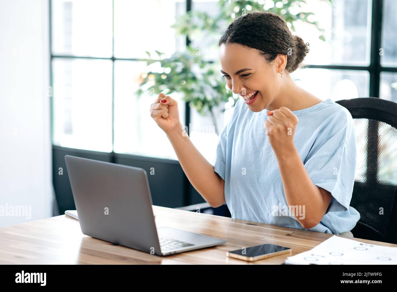 Joyful amazed happy brazilian or hispanic young business woman, sitting at a desk with a laptop in modern office, rejoicing in success, big profit, celebrate deal, gesturing with fists, smiling Stock Photo