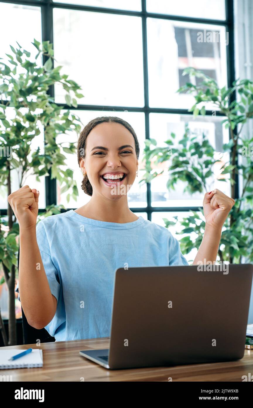 Vertical photo of excited happy mixed race young business woman, sit at a desk with laptop in office, rejoicing in success, big profit, celebrate deal, gesturing with fists, looks at camera, smiling Stock Photo