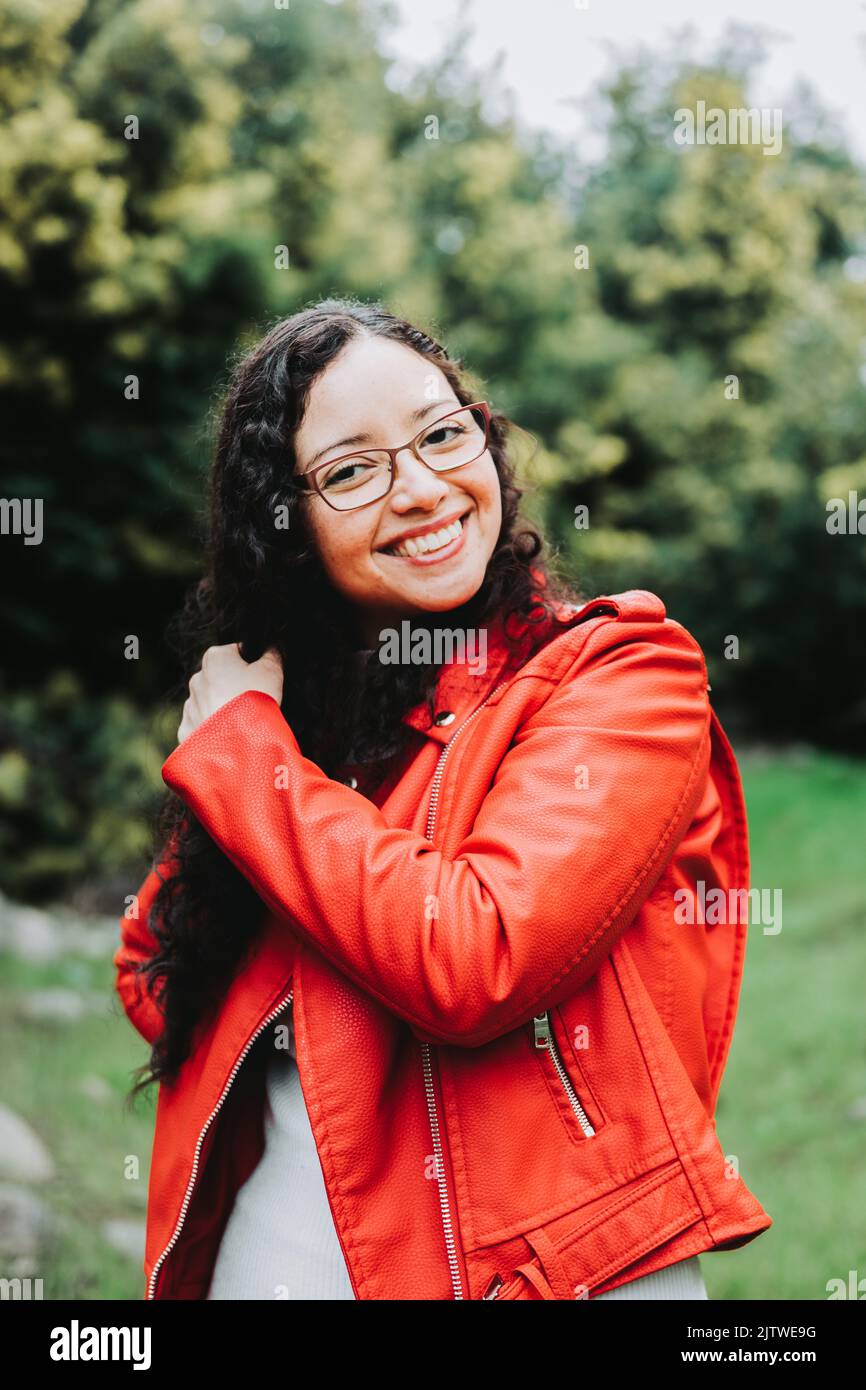Smiling curly brunette woman, wearing a red leather jacket in nature. Stock Photo