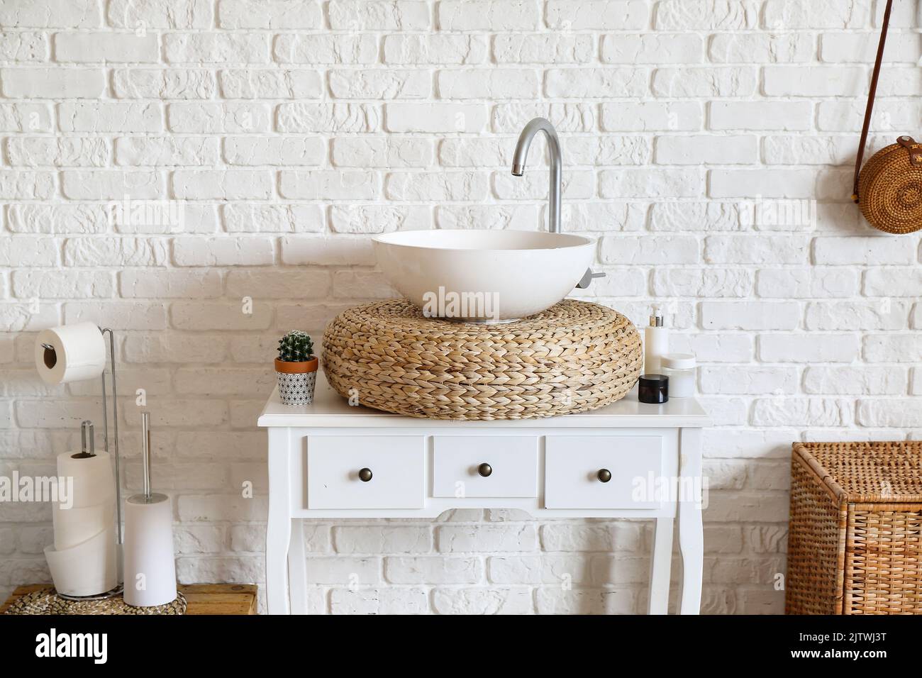 Table with sink, rattan pouf and cosmetic products near white brick wall Stock Photo