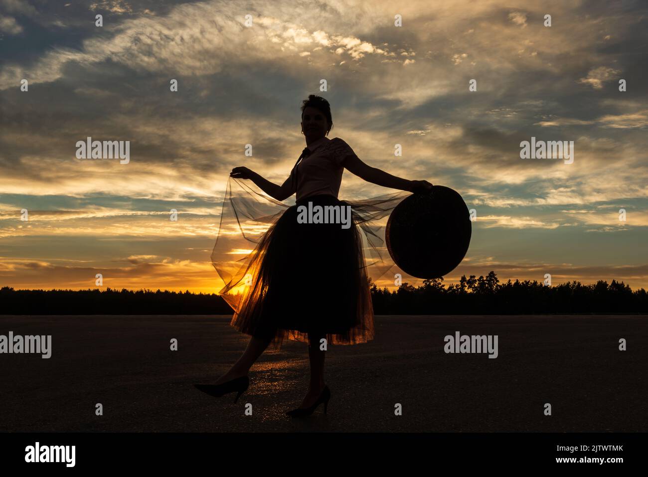 Graceful fairy tale mistress of woodland in backlight of sunset dancing on road with hat in hand against forest skyline and dramatic yellow cloudy sky Stock Photo