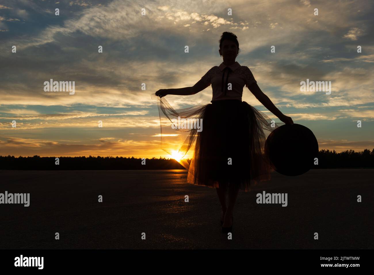 Woman holding edge of tutu skirt against forest silhouette and sunset cloudy sky backlit by sunbeams of setting sun with hat in hand,low angle view Stock Photo