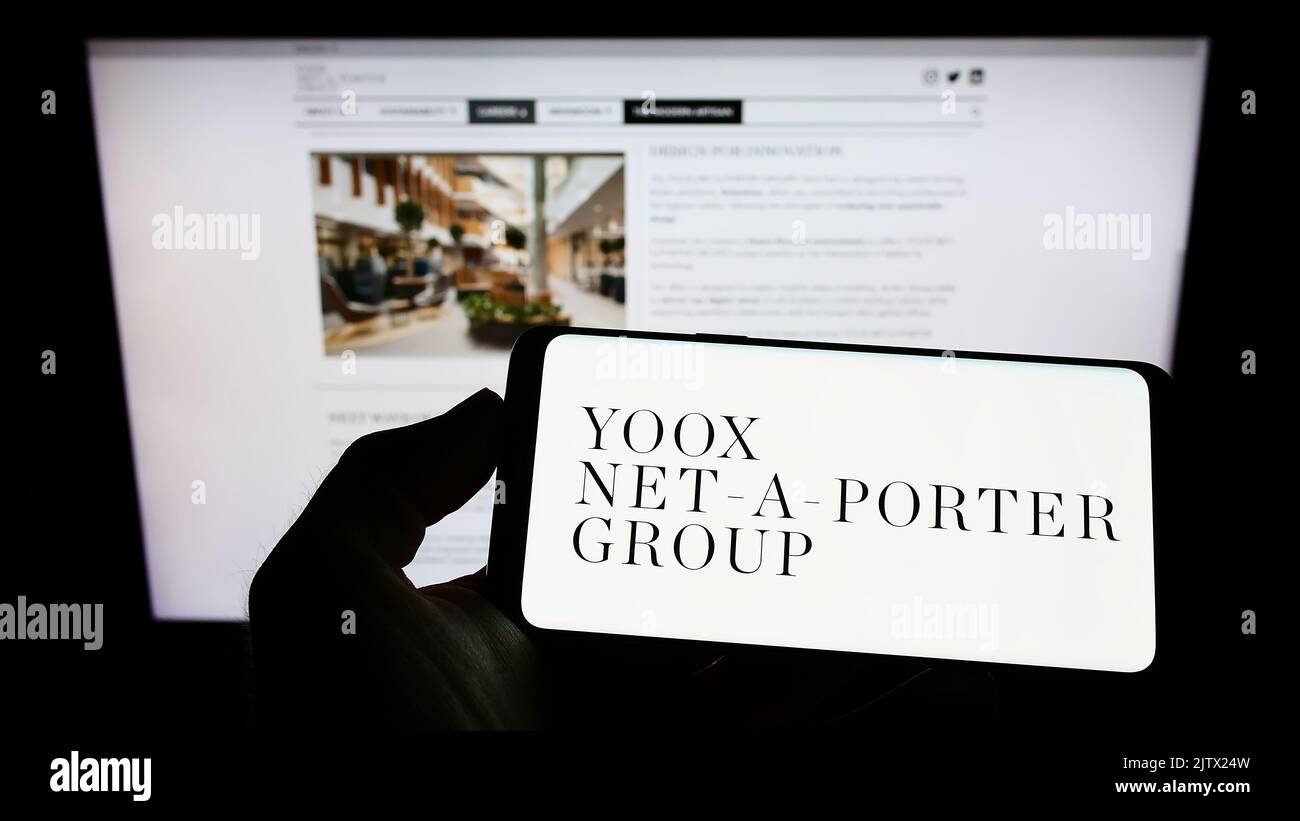 Person holding cellphone with logo of company YOOX Net-a-Porter Group S.p.A. (YNAP) on screen in front of webpage. Focus on phone display. Stock Photo
