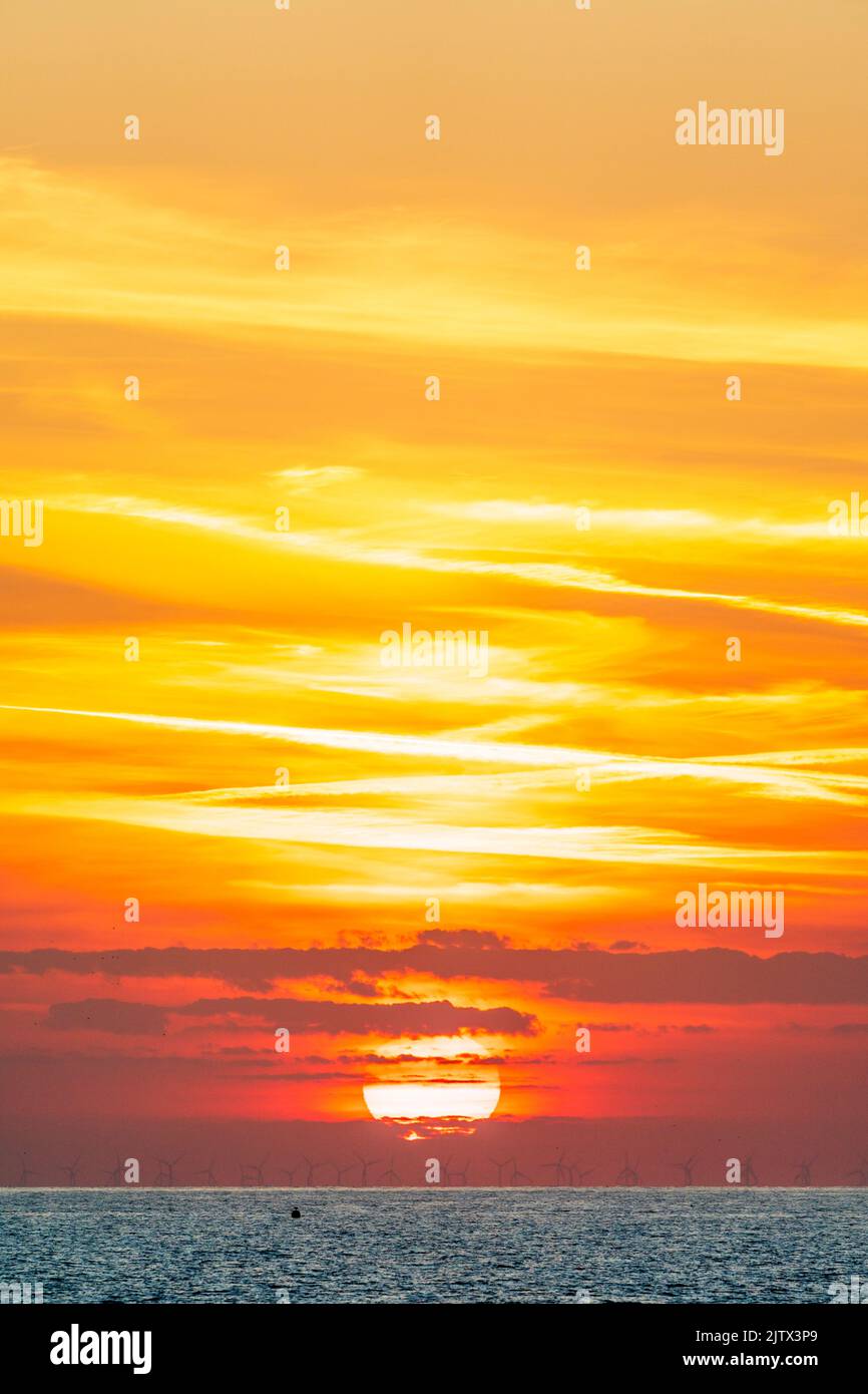 The sun rising into an orange and yellow sky from behind a layer of cloud on the horizon over the North Sea off the Kent coast. Various layer of thin cloud above the sun brightly lit by the sunlight. Stock Photo