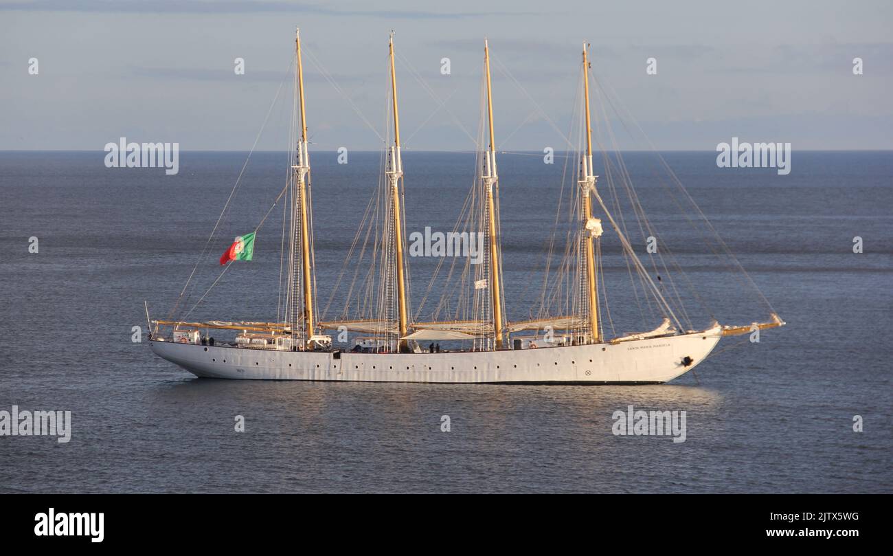 White schooner SANTA MARIA MANUELA, waiving flag of Portugal, anchored off the coast, in sunset light, Angra do Heroismo, Terceira, Azores, Portugal Stock Photo
