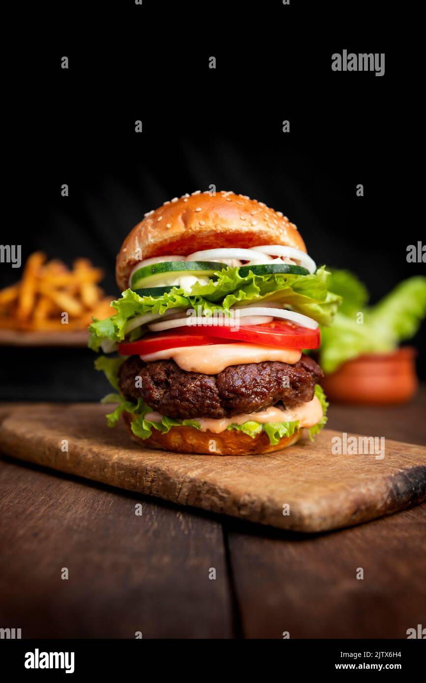 Burger with beef meat, cheese, lettuce, onion, tomato, cucumber, mayo and ketchup on an old wooden table. Junk and unhealthy food concept. Stock Photo