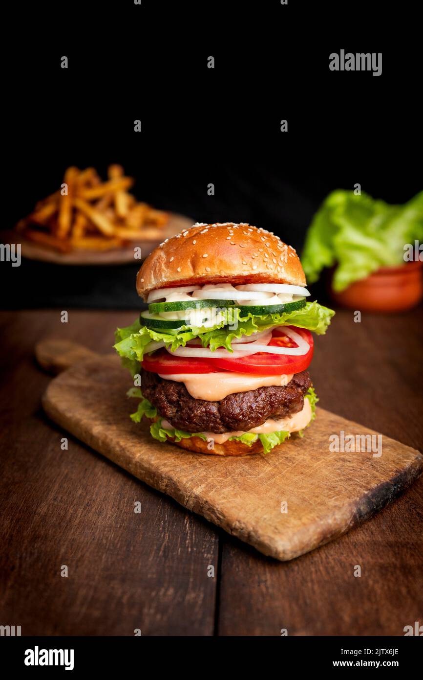 Burger with beef meat, cheese, lettuce, onion, tomato, cucumber, mayo and ketchup on an old wooden table. Junk and unhealthy food concept. Stock Photo