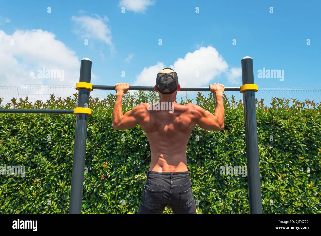 Muscular man in a cap doing pull-ups on the horizontal bar in the park outdoors, back view of his back Stock Photo