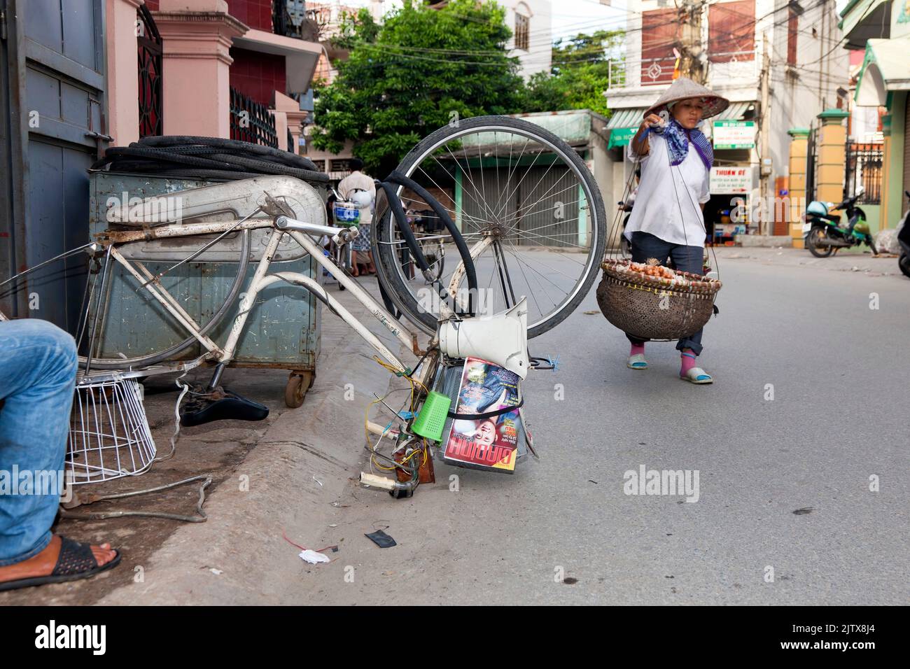 Vietnamese street vendor carrying pannier baskets, and bicycle repair in the road, Hai Phong, Vietnam Stock Photo