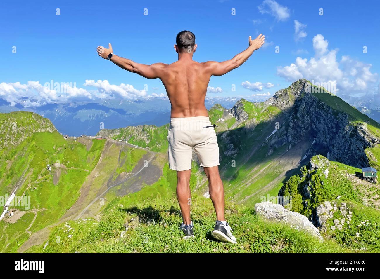 Tanned man with a naked torso in shorts with his arms raised on a mountain top in summer among alpine meadows a tourist on a rock. Travel concept, mou Stock Photo