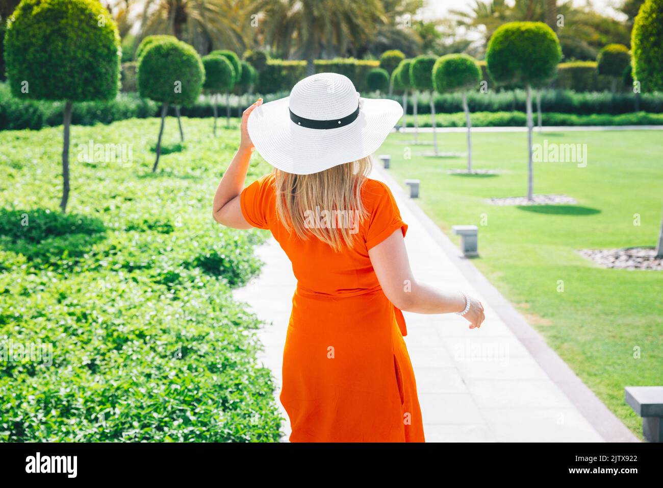 High quality photo. Young Caucasian Woman Lady In Red Dress And White Summer Hat Standing On Path In Zabeel Park And Looking Into Distance At Trees Stock Photo