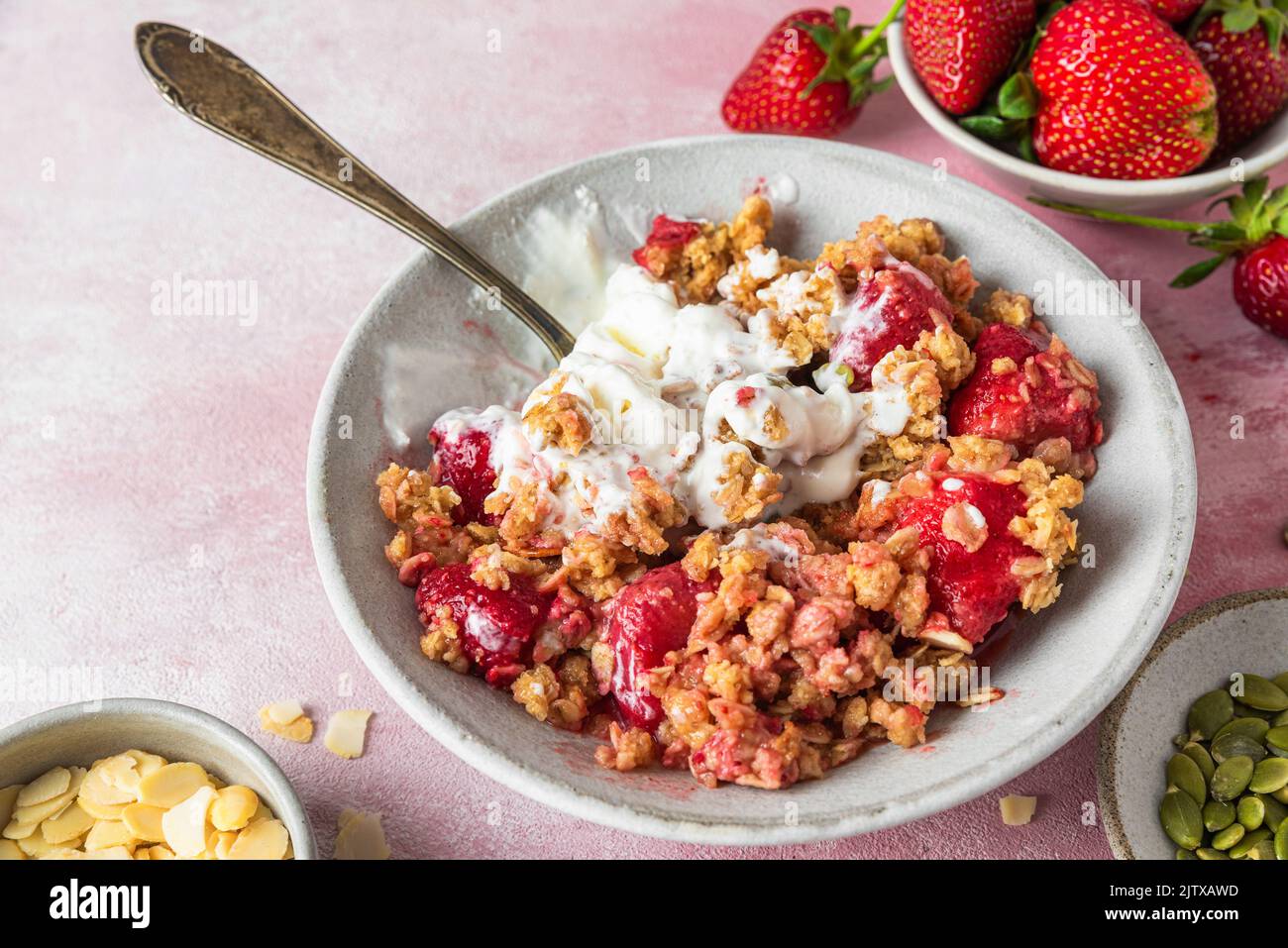Delicious strawberry crumble with ice cream, almonds and pumpkin seeds in a plate with spoon on pink background. Close up. Tasty breakfast Stock Photo