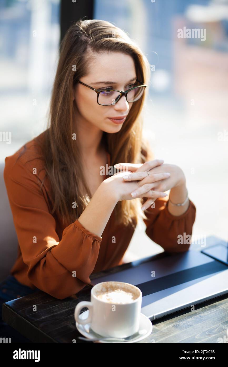 Young businesswoman in a cafe bar or restaurant. Freelancer girl working on laptop and having tea at a window table. Stock Photo