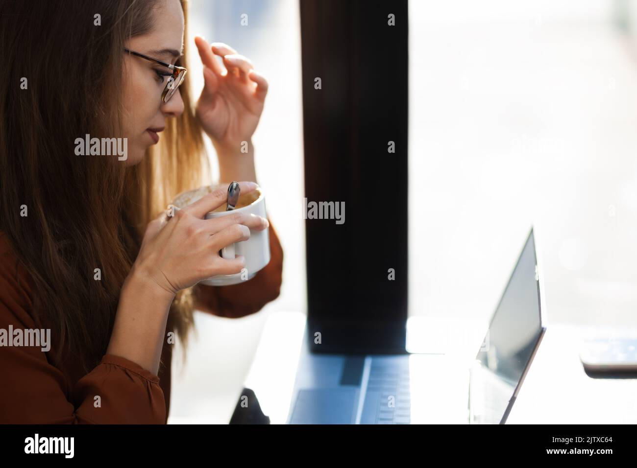 Young businesswoman in a cafe bar or restaurant. Freelancer girl working on laptop and having tea at a window table. Stock Photo