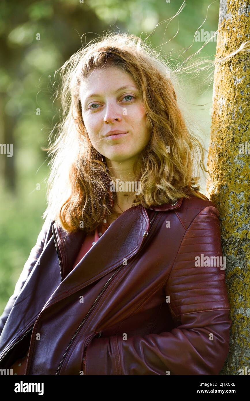Jeune femme portant un blouson de cuir, adossee au tronc d'un arbre. Departement d'Eure-et-Loir, region Centre-Val-de-Loire, France, Europe / Young Stock Photo