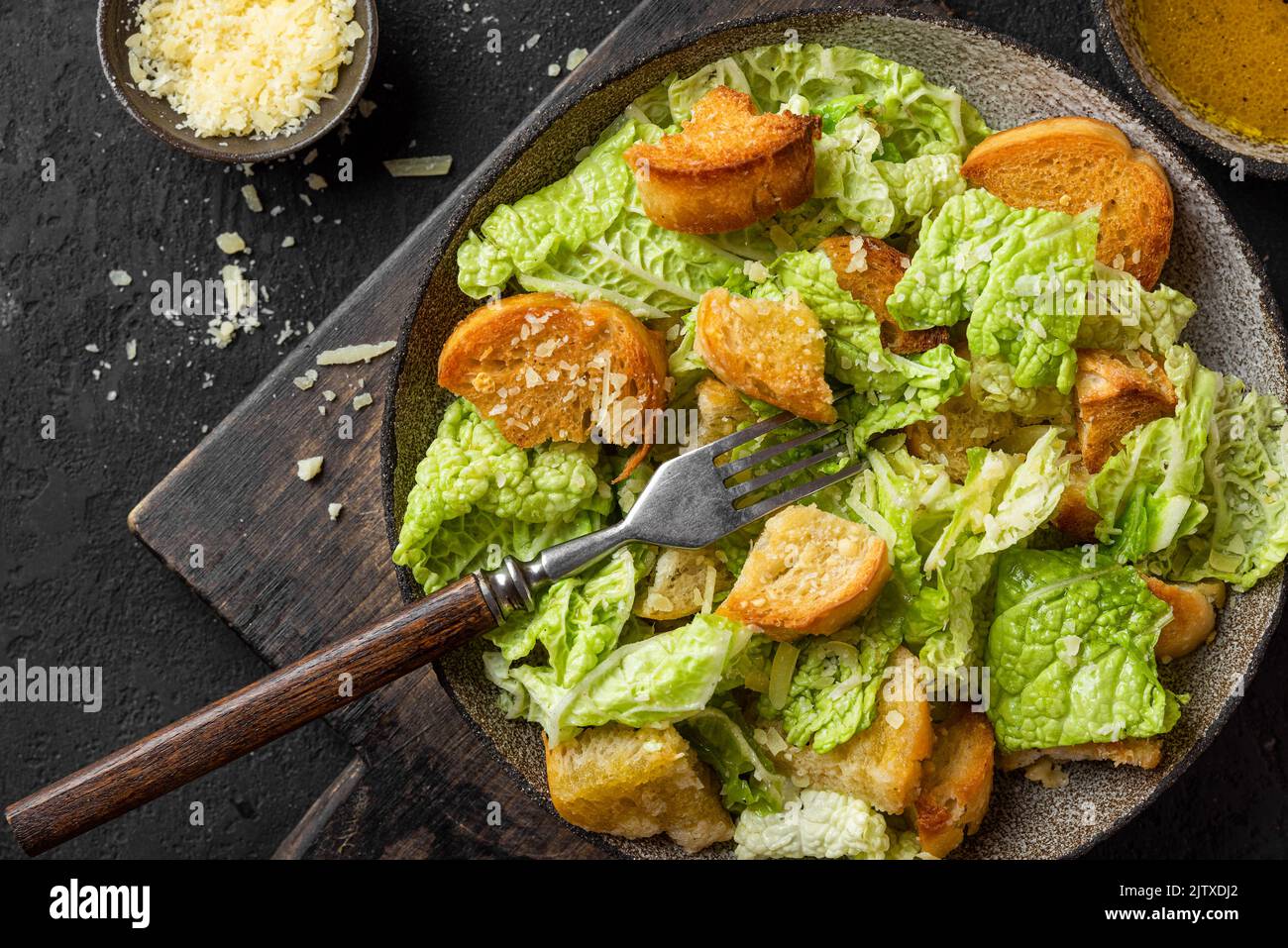 Caesar salad with crisp crotons, parmesan cheese and dressing for tasty lunch or dinner in a plate with fork on black background. Top view Stock Photo