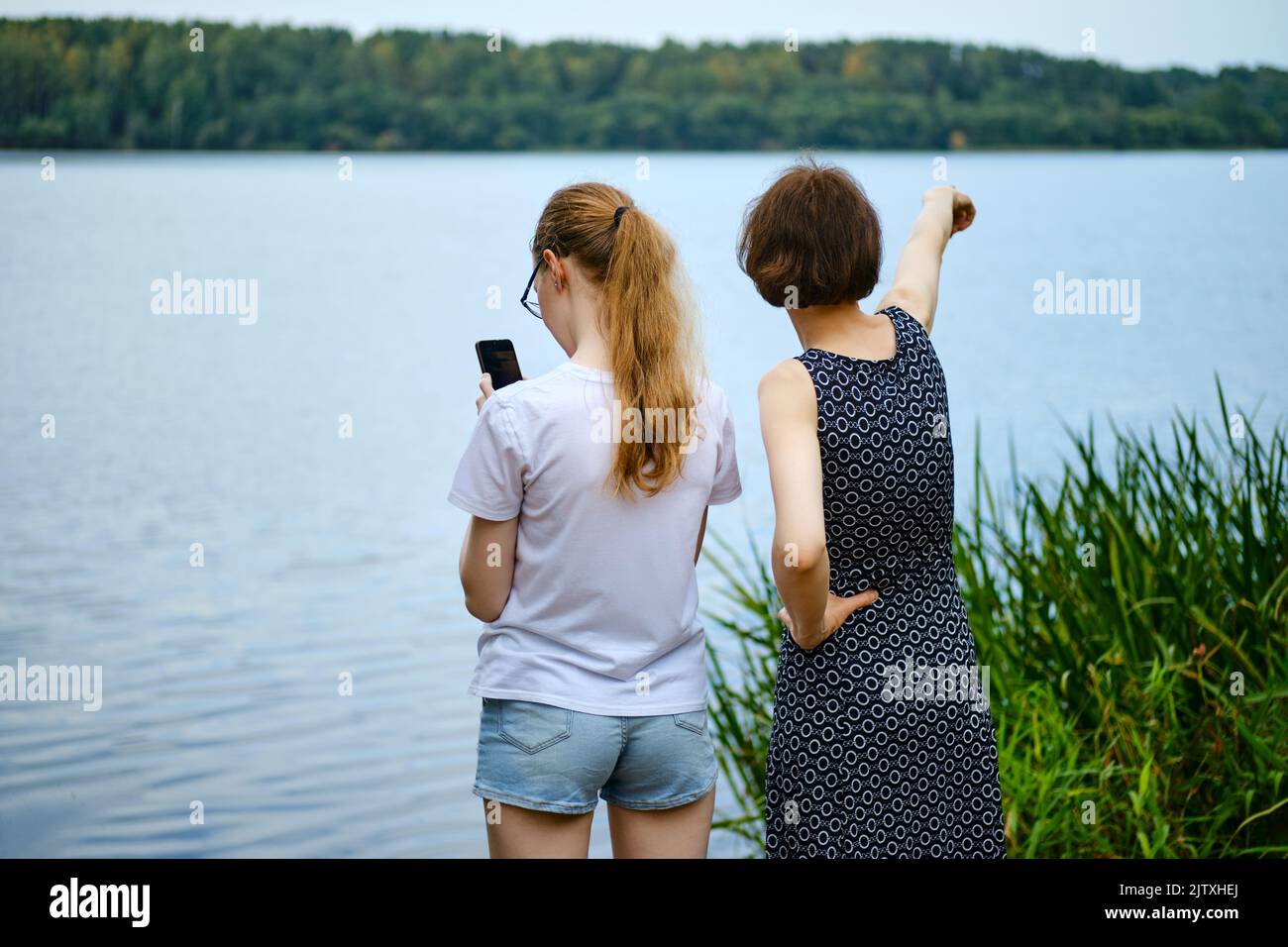 Mother shows to her daughter something far away at the shore of the lake and daughter plots a route on smartphone Stock Photo