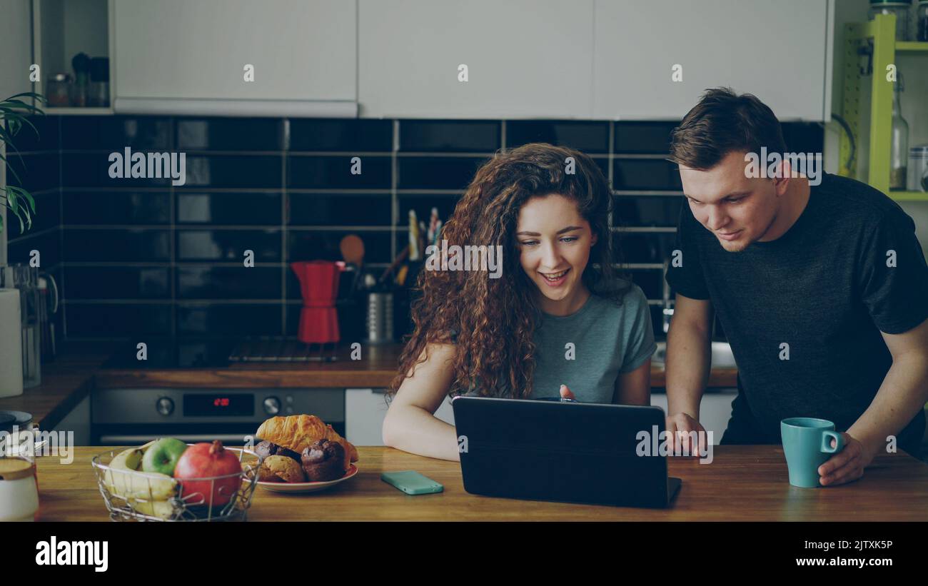 Young positive caucasian couple sitting at table in kitchen using laptop smiling and discussing at home Stock Photo