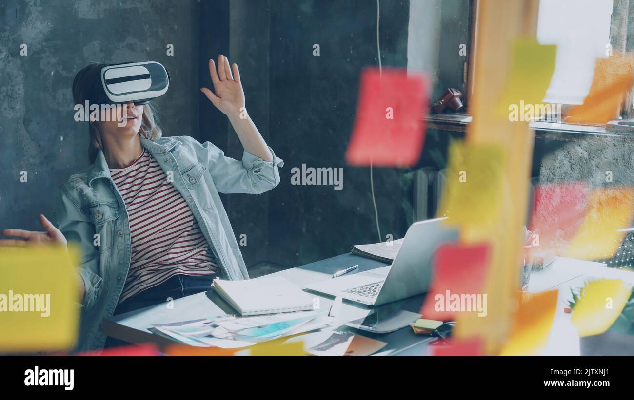 Attractive young woman is sitting at table and wearing virtual reality glasses. She is moving hands and and having VR experience in modern lof office indoors Stock Photo