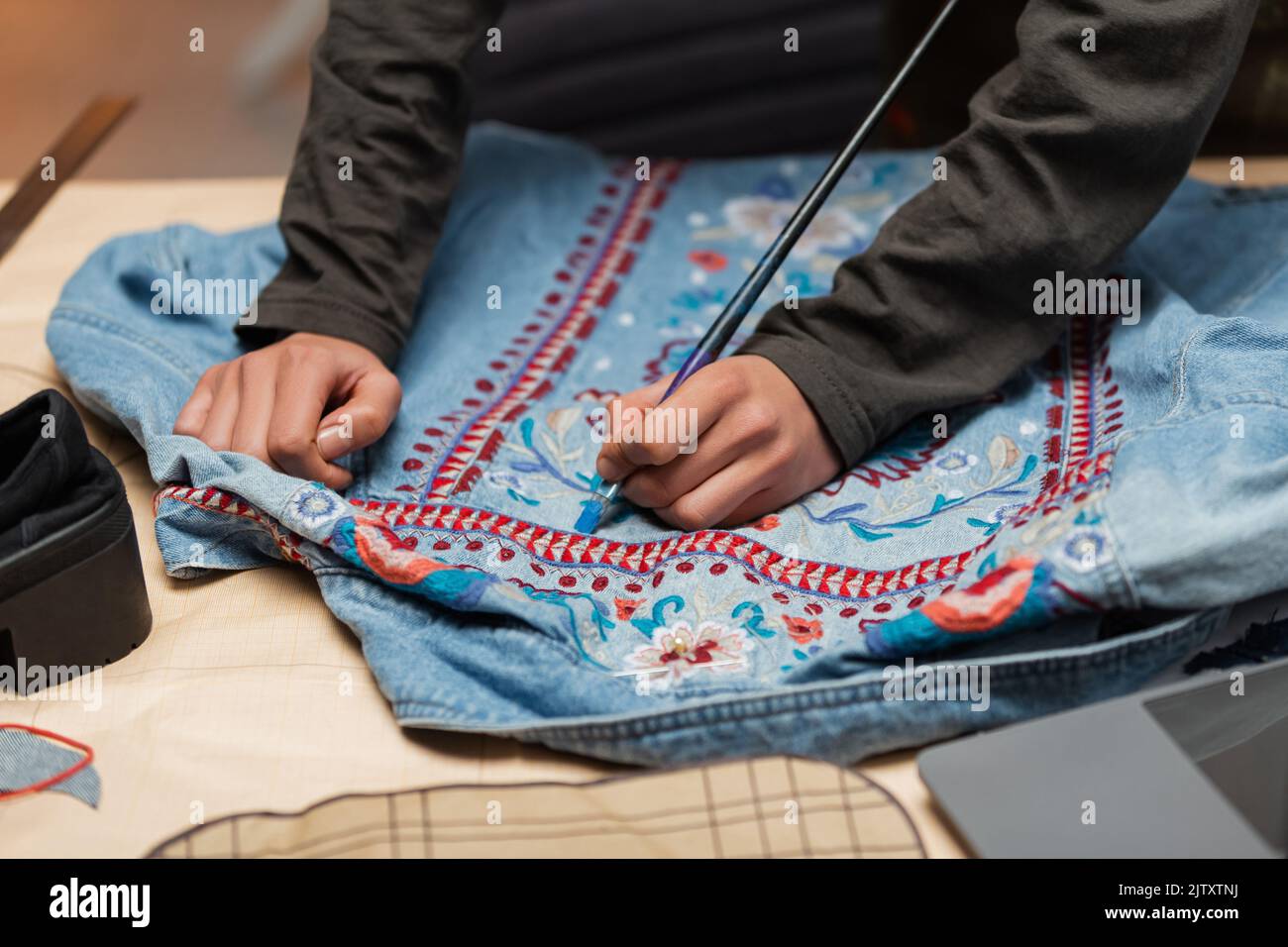 Cropped view of african american craftswoman painting on denim jacket near blurred laptop in workshop,stock image Stock Photo