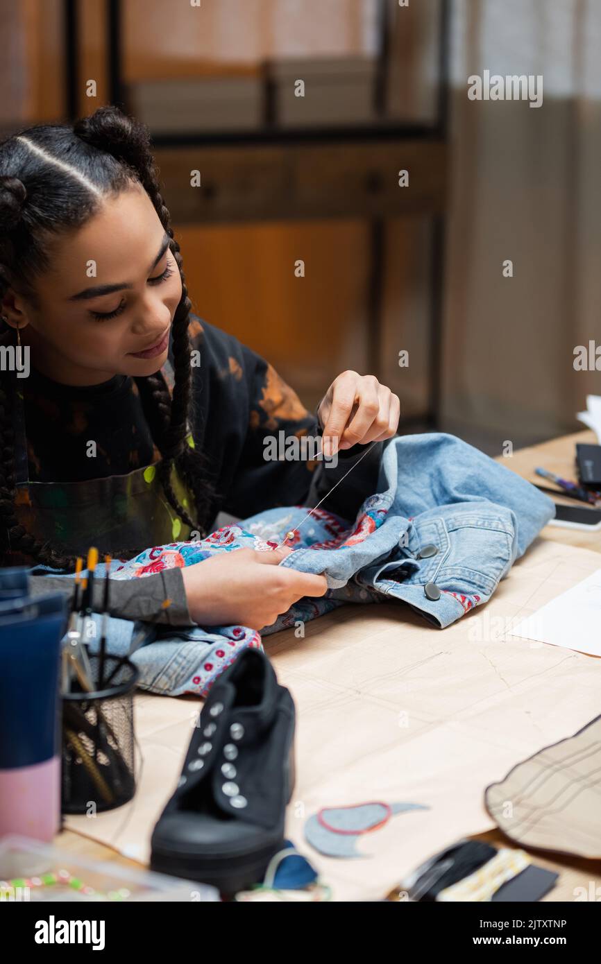 Smiling african american craftswoman embroidering denim jacket near sewing pattern in workshop,stock image Stock Photo