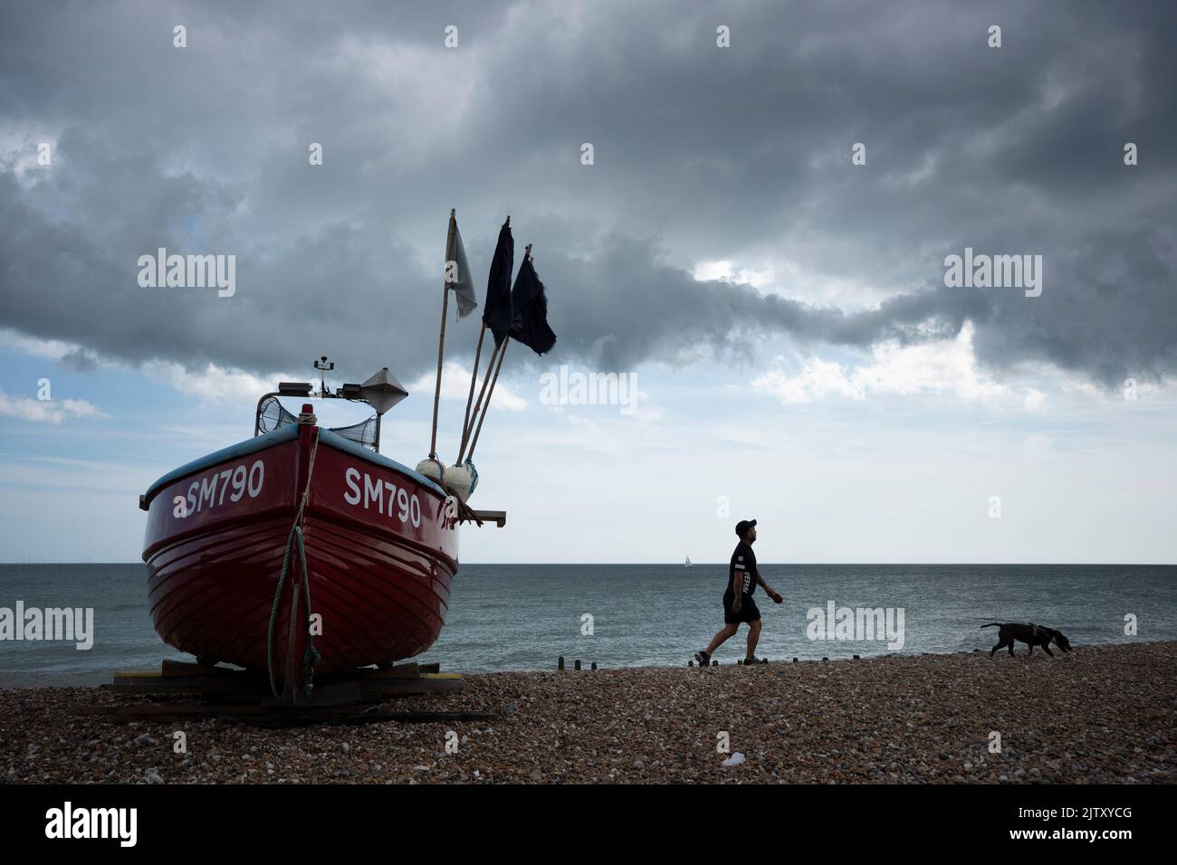 A man walks past a beached fishing boat on the shingle beach at Worthing, on 29th August 2022, in Worthing, England. Stock Photo