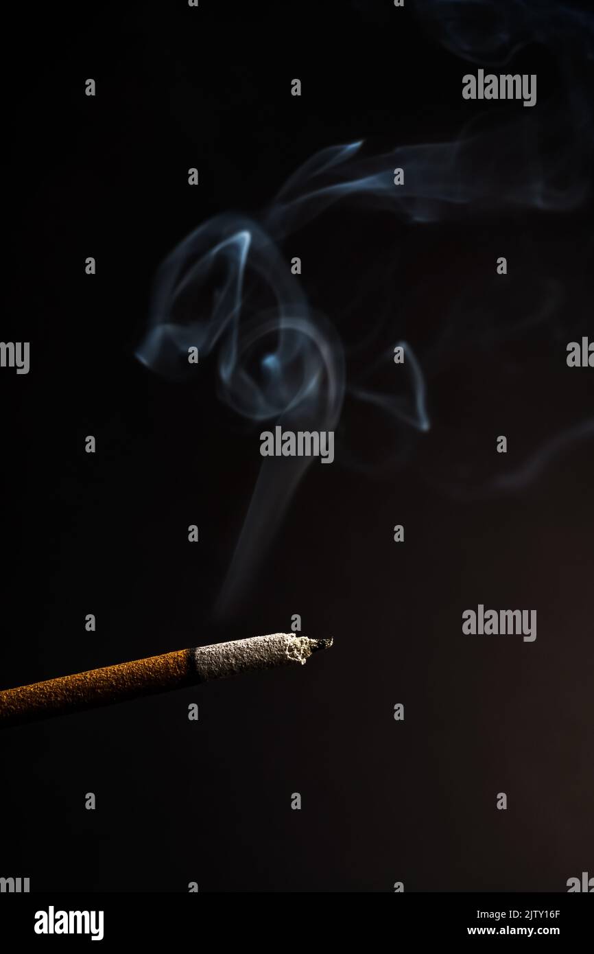 close-up of a burning incense with white smoke coming out illuminated on a black background Stock Photo