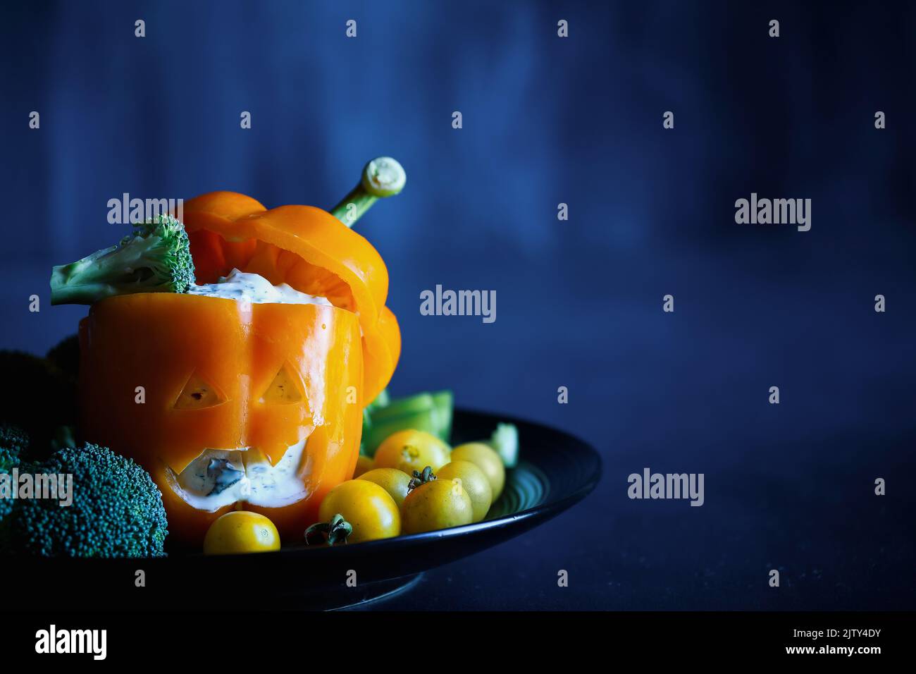 Colorful stuffed sweet orange bell pepper as monster with cut out face carved into a Halloween pumpkin Jack O'Lantern and filled with Tzatziki dip. Se Stock Photo