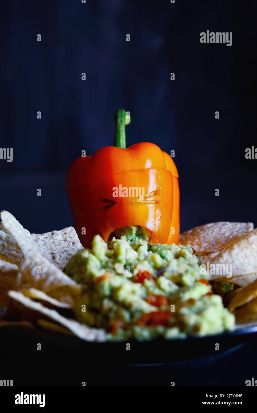 Colorful stuffed sweet orange bell pepper as monster with cut out face carved into a Halloween pumpkin Jack O'Lantern throwing up guacamole Stock Photo