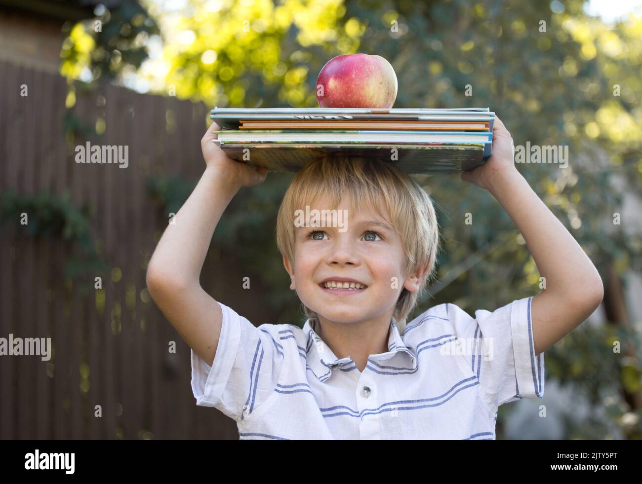 cute joyful 6 year old boy is holding a stack of books and a big apple on his head. Reading books, interesting childhood, inquisitive child, knowledge Stock Photo