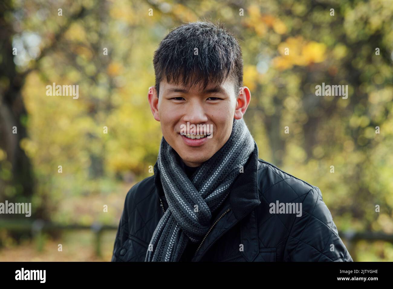 A portrait of a young, Chinese man standing outdoors in nature in Northumberland, North East England during Autumn. He is smiling while looking at the Stock Photo