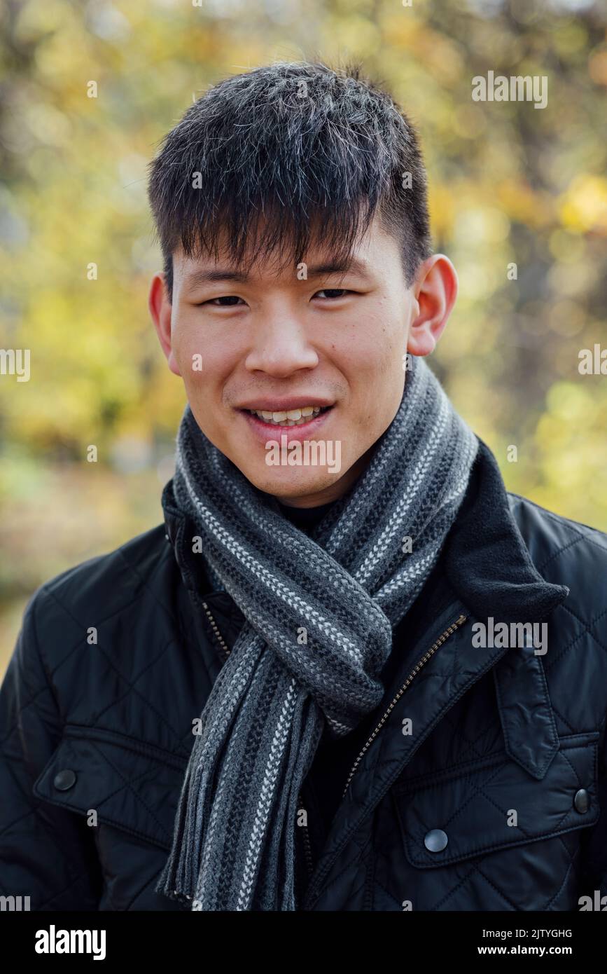 A close-up portrait of a young, Chinese man standing outdoors in nature in Northumberland, North East England during Autumn. He is smiling while looki Stock Photo