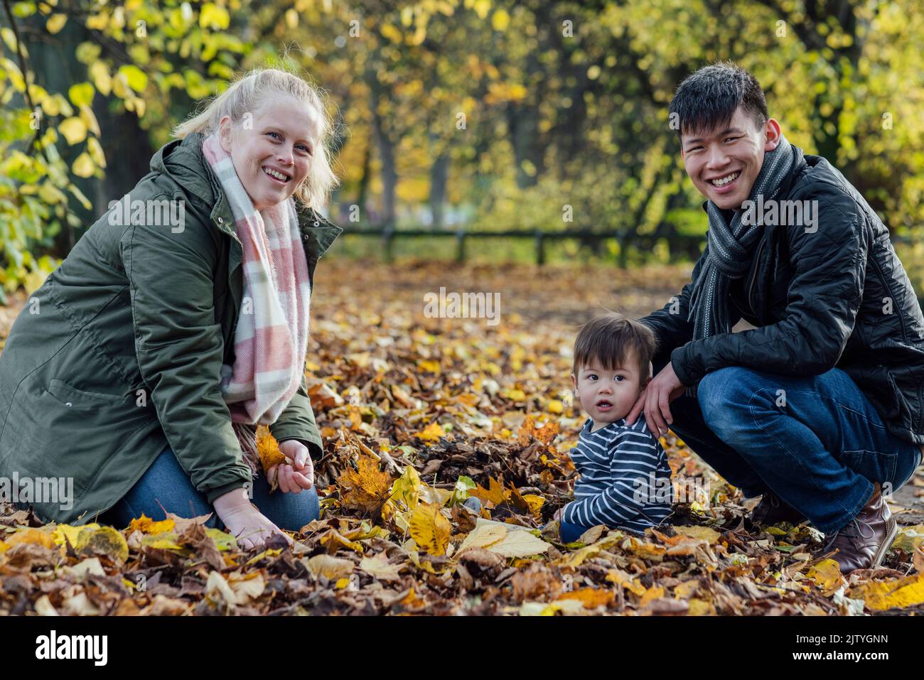 A family crouching down and playing with fallen leaves in nature in Northumberland, North East England. They are all looking at the camera and smiling Stock Photo