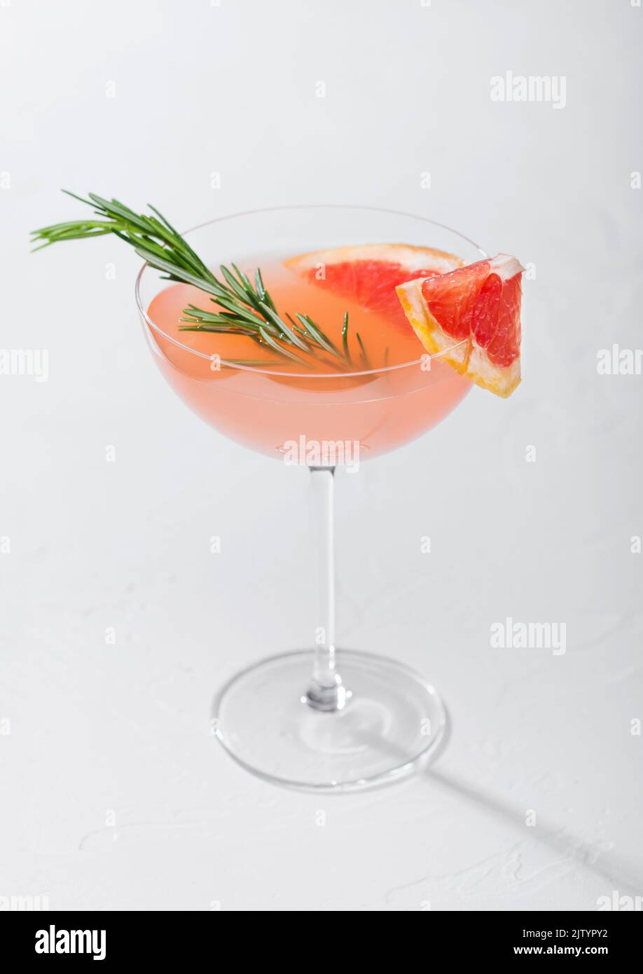 Red grapefruit summer cocktail in crystal martini glass with fruit slice and rosemary on white background Stock Photo