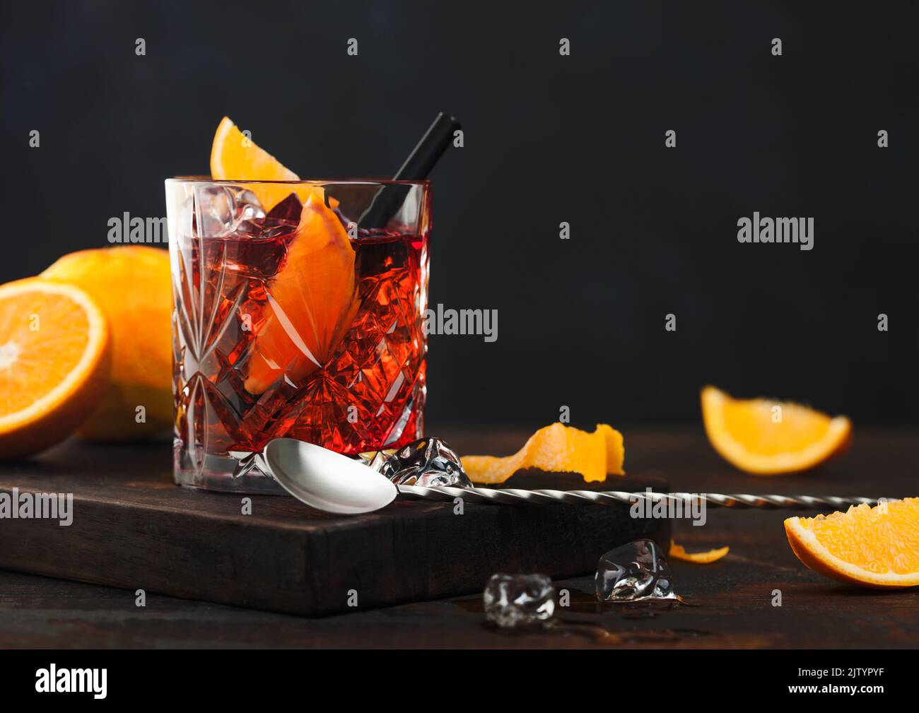Negroni cocktail in crystal glass with orange slice and fresh raw oranges on chopping board with spoon on wooden background. Stock Photo