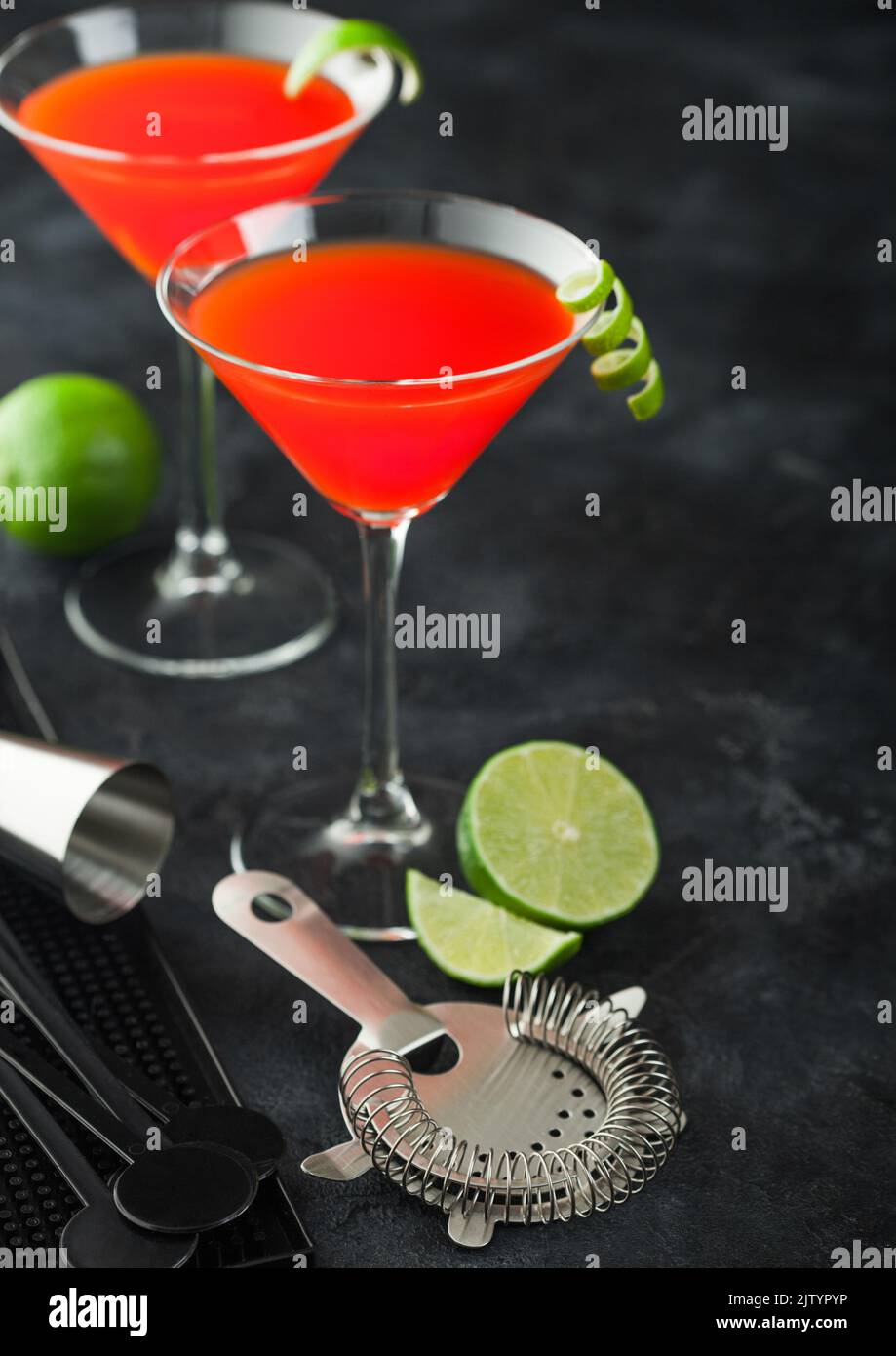 Cosmopolitan cocktail in classic crystal glasses with lime peel and fresh limes with strainer on black table background. Top view Stock Photo