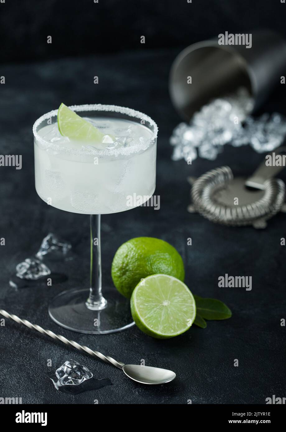 Luxury glass of Margarita cocktail with fresh limes and bar spoon with ice cubes in shaker on black table background. Stock Photo