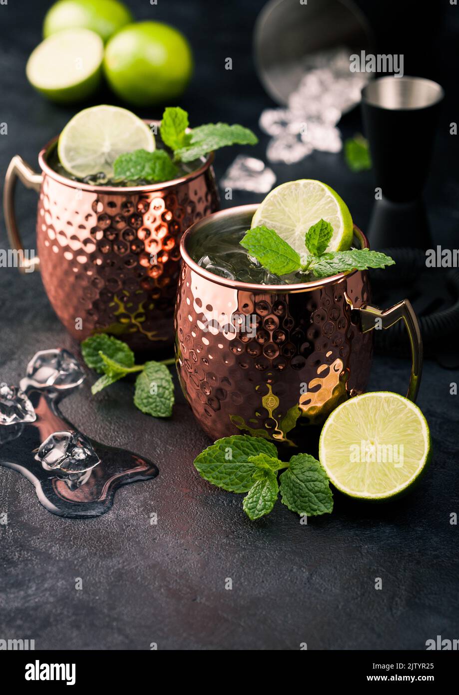Moscow mule cocktail in a copper mug with lime and mint and wooden squeezer on dark kitchen background with ice cubes. Stock Photo