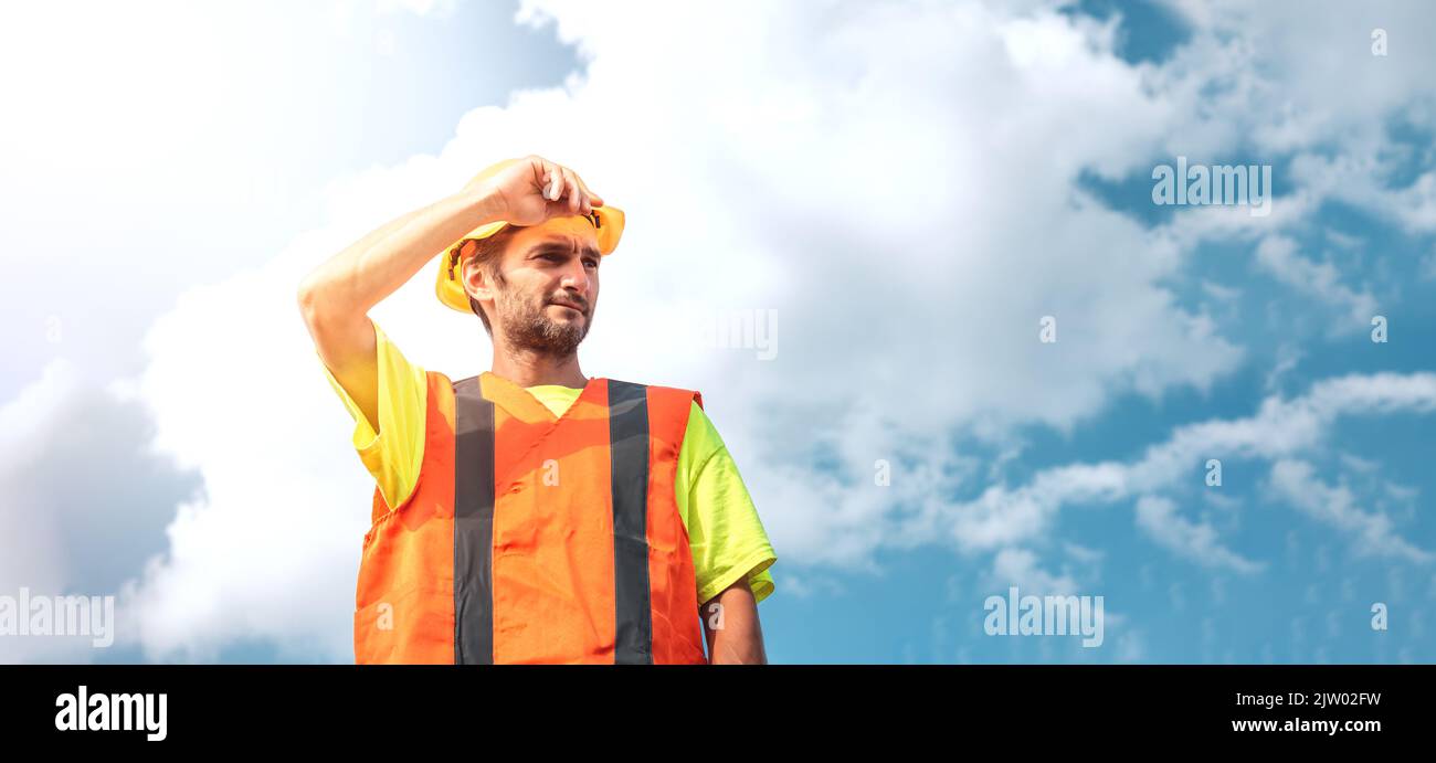 A portrait of a worker stands with confidence in an orange work suit and safety helmet against the sky. Smart industrial worker work concept. Stock Photo
