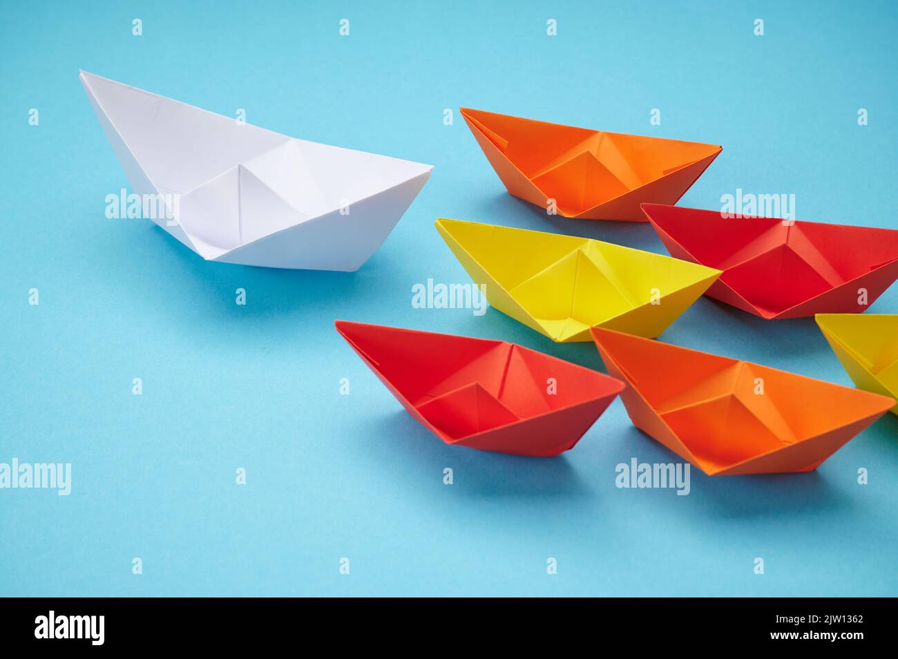 white paper boat leads a group of colored paper boats on blue background Stock Photo
